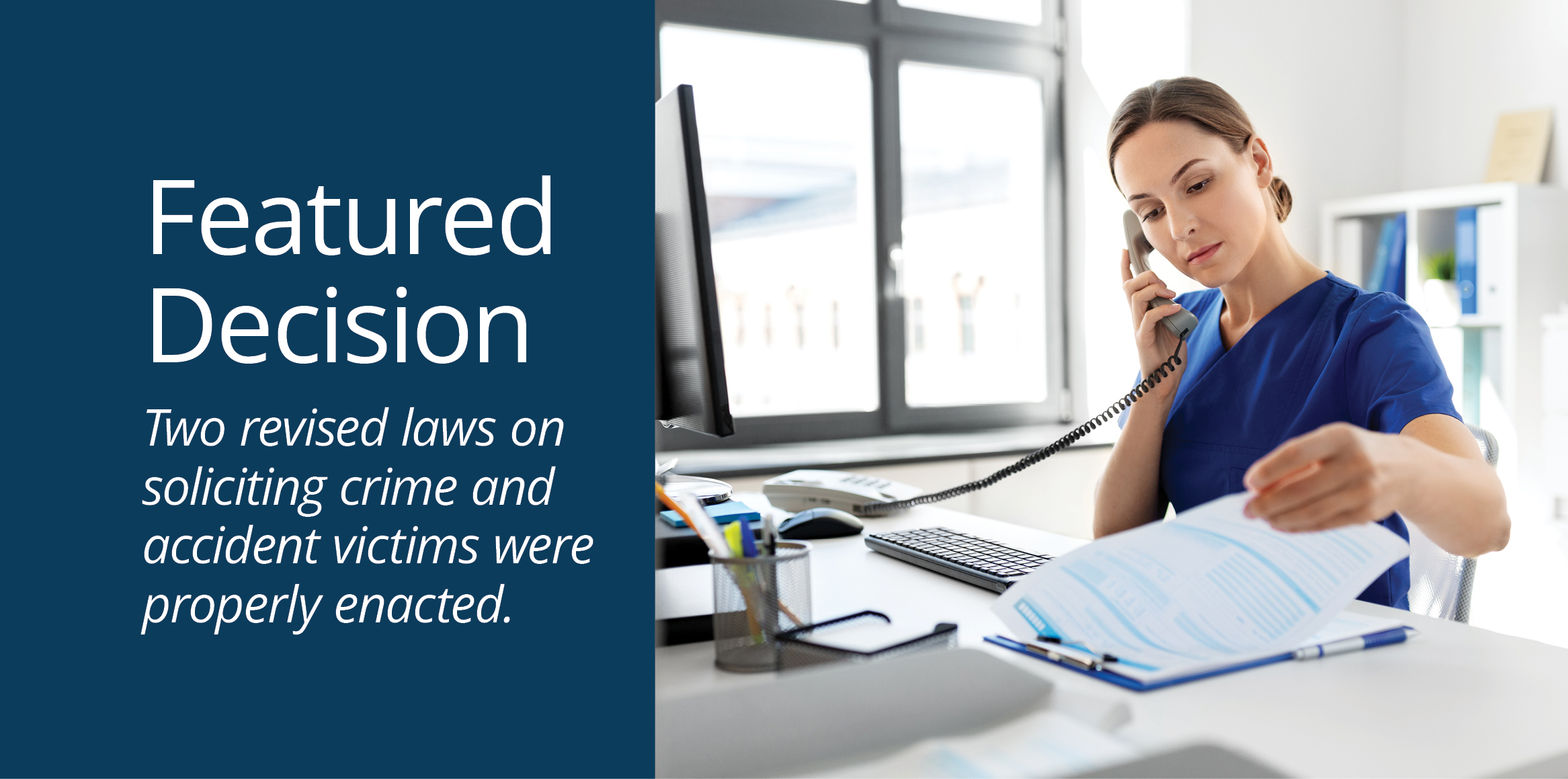 An administrative healthcare worker in blue scrubs sits at a bright office desk, speaking on a landline phone while reviewing a document on a clipboard.